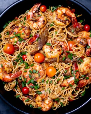 Spaghetti pasta with shrimp and cherry tomatoes in a black pan garnished with spring onions.