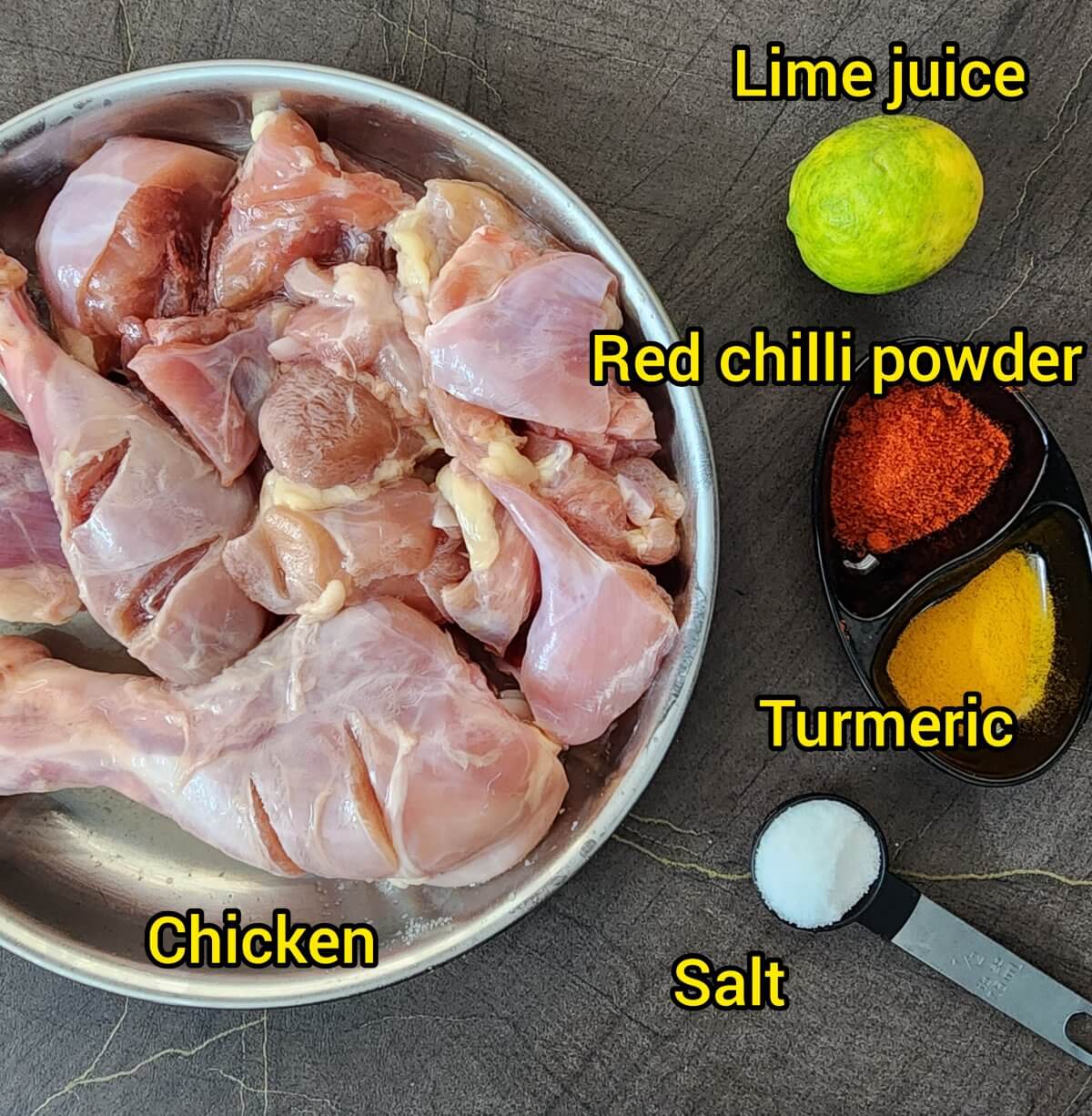 Chicken thighs and drumsticks in a steel plate. Salt in spoon measure. Red Chilli and turmeric powders in black bowl. Lemon.