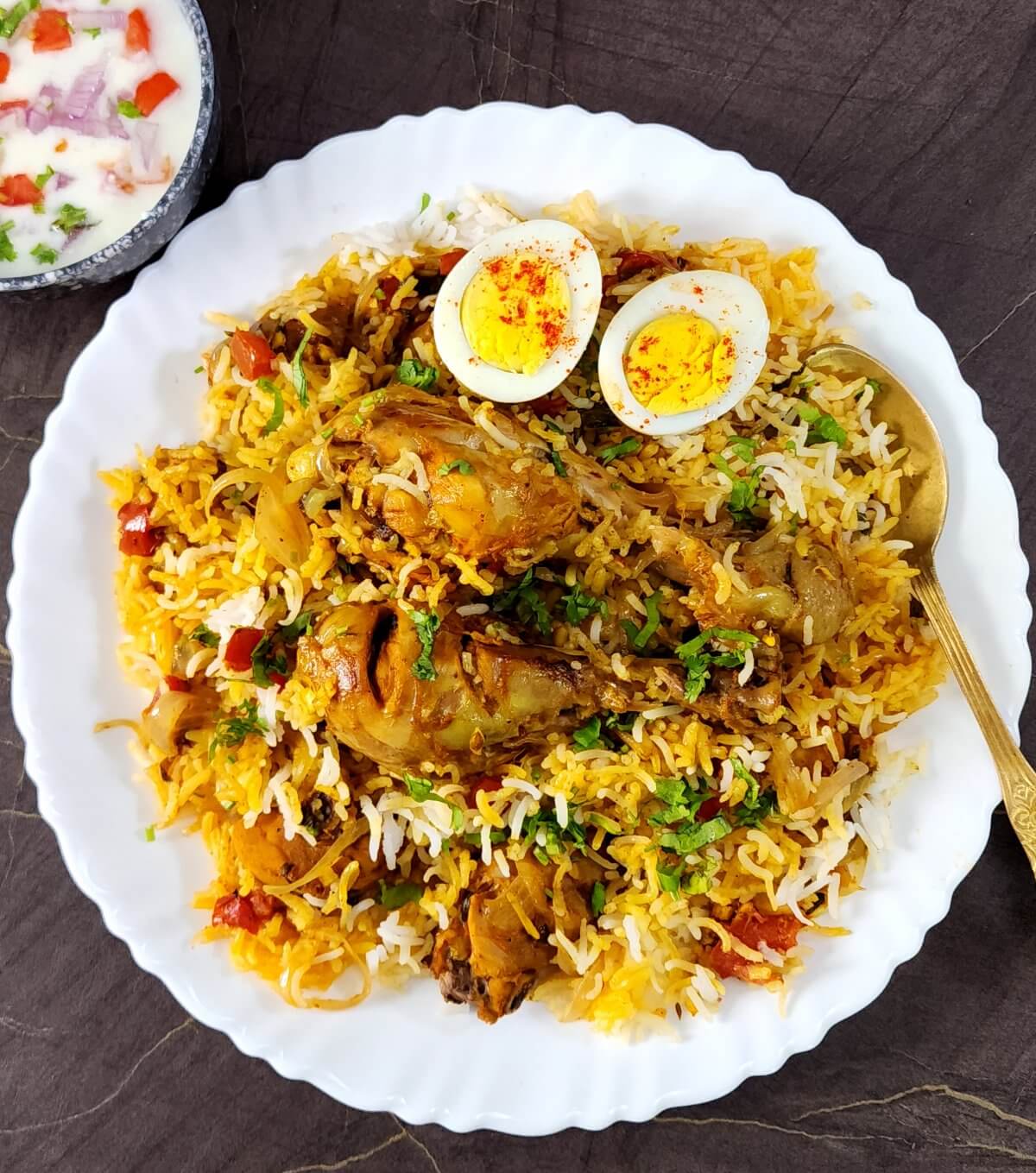 Authentic chicken biryani served in a white plate along with golden spoon and a bowl of raita/yogurt salad.