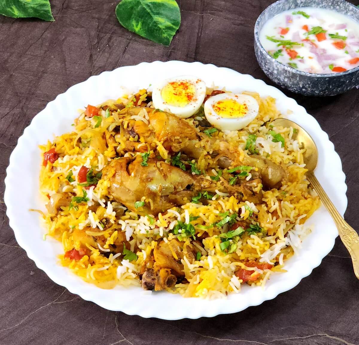 Hyderabadi Chicken Biryani served in a white plate along with boiled eggs and raita.