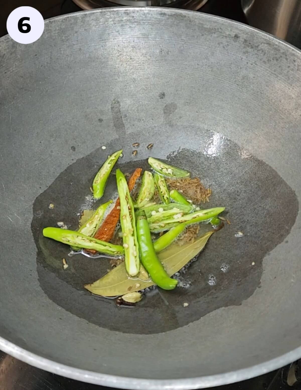 Whole garam masala and slit green chilies sizzling in oil within a large pan/kadhai.